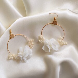 May include: A pair of gold hoop earrings with white fabric flowers and small dried flowers. The earrings have a gold hook and a small gold disc at the top of the hoop. The earrings are displayed on a white satin background.
