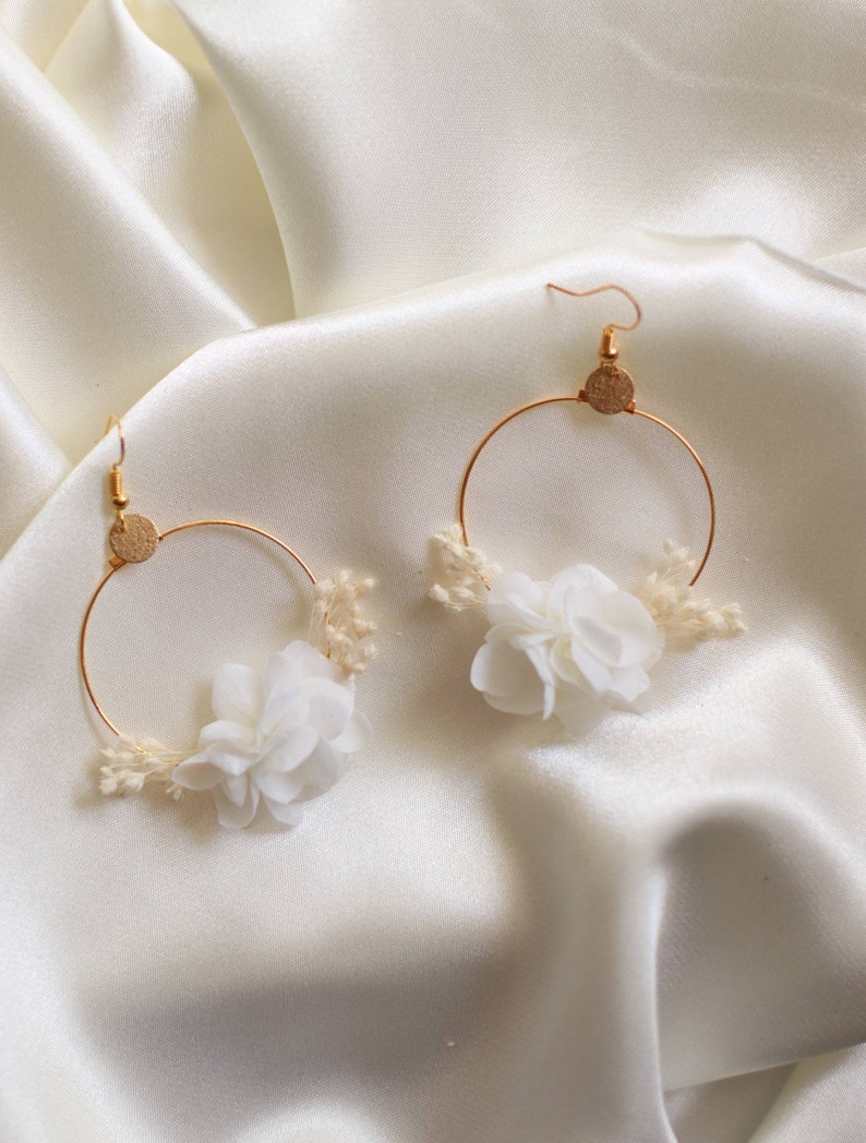 May include: A pair of gold hoop earrings with white fabric flowers and small dried flowers. The earrings have a gold hook and a small gold disc at the top of the hoop. The earrings are displayed on a white satin background.