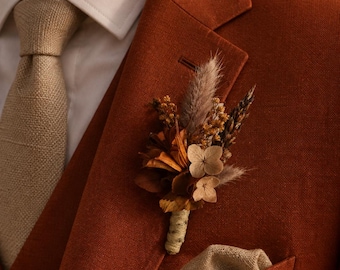 Peut inclure: Une boutonnière composée de fleurs séchées, incluant des pétales d'hortensia beige, des plumets duveteux et de petites fleurs jaunes, le tout entouré de ficelle. Elle est épinglée sur un blazer rouille porté sur une chemise blanche et une cravate beige.