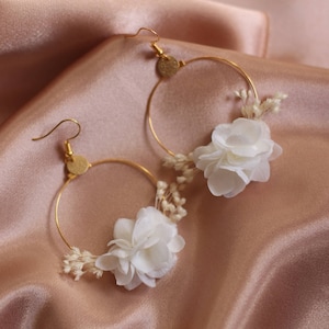 May include: A pair of gold-tone hoop earrings. Each earring features a white flower and small, dried white flowers. The earrings are set against a backdrop of a peach-colored satin fabric.