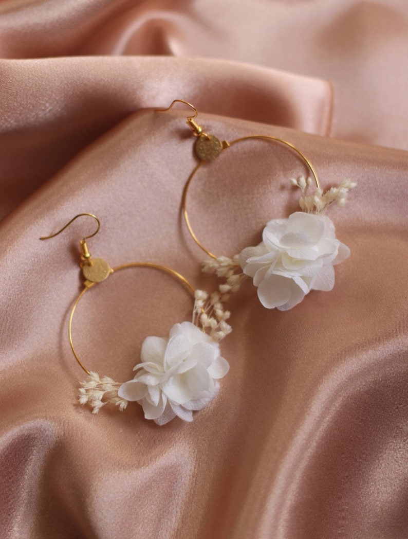 May include: A pair of gold-tone hoop earrings. Each earring features a white flower and small, dried white flowers. The earrings are set against a backdrop of a peach-colored satin fabric.