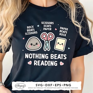 Puede incluir: Camiseta negra con un diseño lúdico que presenta una piedra, unas tijeras y papel, con el texto "NADA SUPERA LA LECTURA". El diseño incluye las frases "Piedra vence a tijeras", "Tijeras vencen a papel" y "Papel vence a piedra".