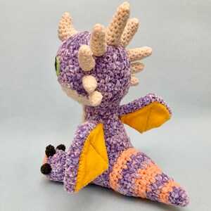 Peut inclure: Peluche de dragon en crochet violet et orange avec des ailes en feutre jaune. Le dragon a une corne en crochet blanc et des yeux verts.