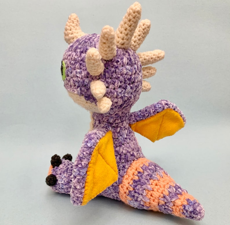 Peut inclure: Peluche de dragon en crochet violet et orange avec des ailes en feutre jaune. Le dragon a une corne en crochet blanc et des yeux verts.