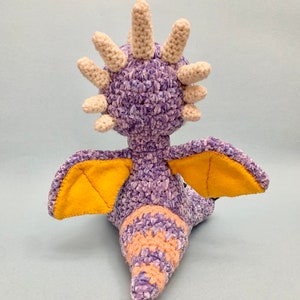 Peut inclure: Jouet en crochet de dragon violet et jaune avec une cr&ecirc;te blanche h&eacute;riss&eacute;e. Le dragon a des ailes et une queue.