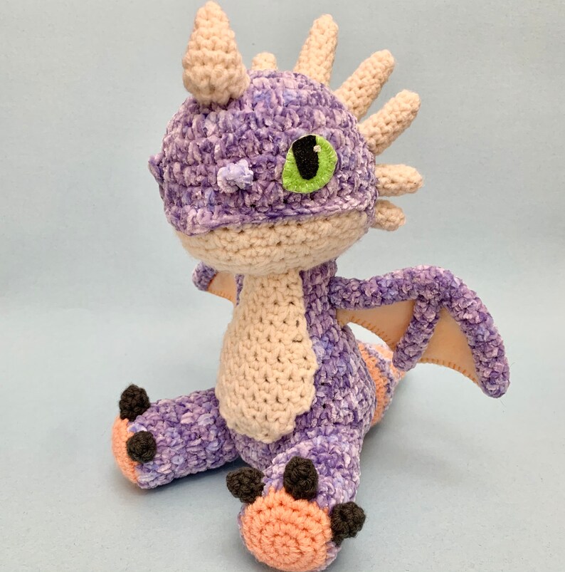 Peut inclure: Peluche de dragon en crochet violet et cr&egrave;me avec un &oelig;il vert. Le dragon a une t&ecirc;te h&eacute;riss&eacute;e et des ailes.
