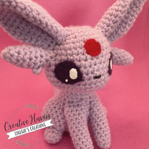 May include: A crocheted lavender-colored plush toy with large ears, a red spot on its forehead, and purple eyes with white accents. The toy is sitting on a pink background. The text "Creative Haven Lindsay's Creations" is in a circular logo.