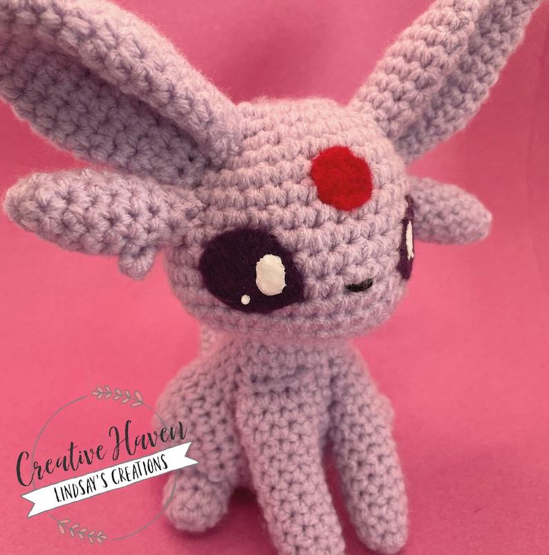 May include: A crocheted lavender-colored plush toy with large ears, a red spot on its forehead, and purple eyes with white accents. The toy is sitting on a pink background. The text "Creative Haven Lindsay's Creations" is in a circular logo.