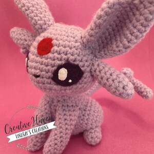 May include: A handmade crochet Espeon plushie in shades of lavender, with large ears, a red spot on its forehead, and white accents around its eyes. The plushie is sitting on a pink surface. The text "Creative Haven Lindsay's Creations" is in the lower left corner.