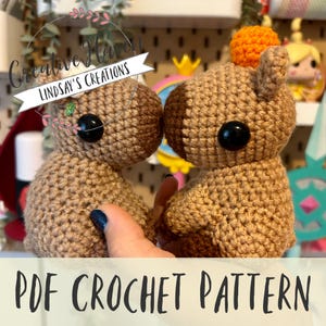 May include: A crochet pattern for two brown amigurumi capybaras with black eyes and orange pom-poms. The pattern is available as a PDF download.  Creative Haven Lindsay's Creations.