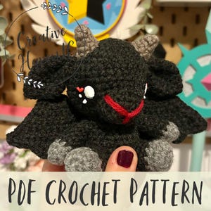 May include: A black crochet amigurumi pattern for a small dragon with red eyes and a red mouth. The dragon has gray feet and wings. The pattern is available as a PDF download.