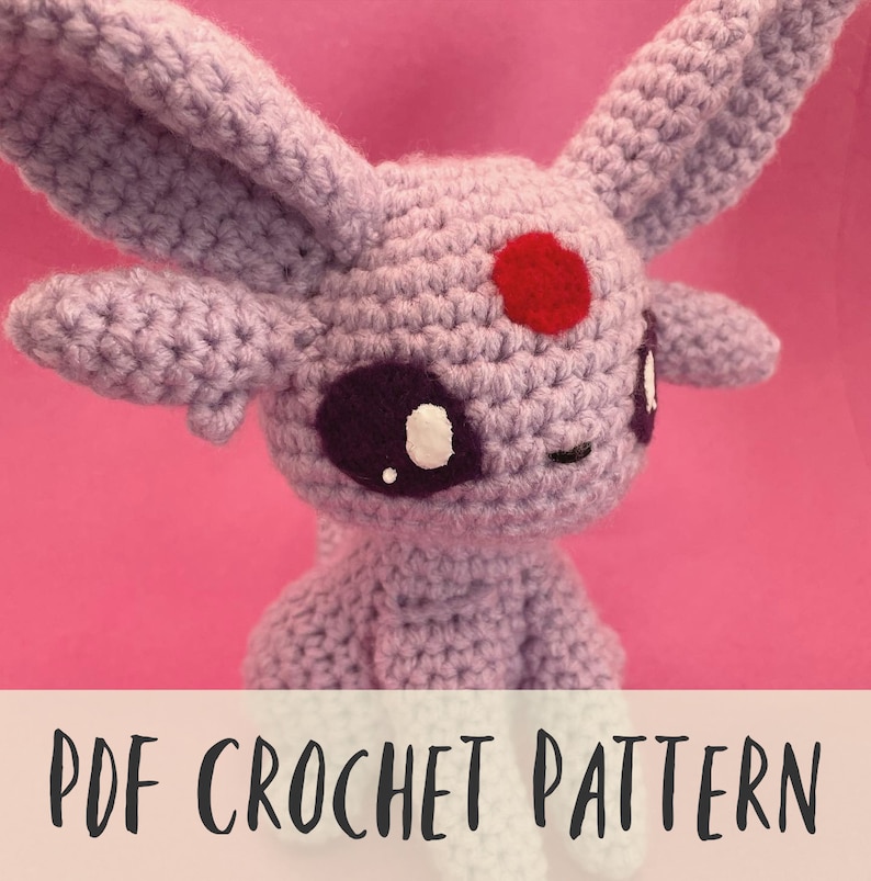 May include: A crocheted lavender-colored plush toy with large ears, a red spot on its forehead, and purple and white eyes. The text "PDF CROCHET PATTERN" is displayed at the bottom of the image. The toy is set against a pink background.