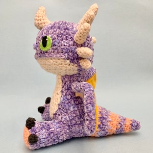 Peut inclure: Peluche de dragon en crochet violet et jaune avec un &oelig;il vert. Le dragon est assis avec sa queue enroul&eacute;e autour de son corps.