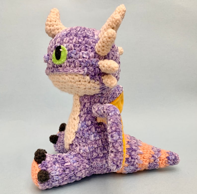 Peut inclure: Peluche de dragon en crochet violet et jaune avec un &oelig;il vert. Le dragon est assis avec sa queue enroul&eacute;e autour de son corps.