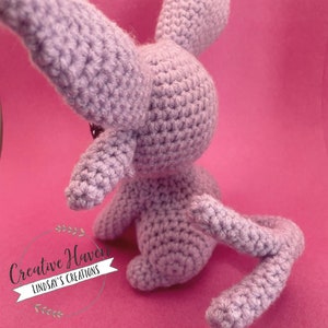 May include: A lavender-colored, crocheted bunny toy with long ears and a curled tail. The handmade stuffed animal is positioned on a pink background. The bunny's body is detailed with visible crochet stitches.