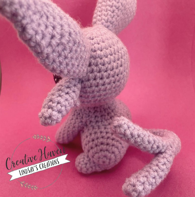 May include: A lavender-colored, crocheted bunny toy with long ears and a curled tail. The handmade stuffed animal is positioned on a pink background. The bunny's body is detailed with visible crochet stitches.