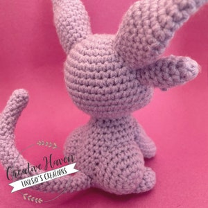 May include: A lavender-colored, crocheted bunny toy with long ears and a curved tail. The handmade plush animal is sitting on a pink background. The text "Creative Haven Lindsay's Creations" is in the lower left corner.