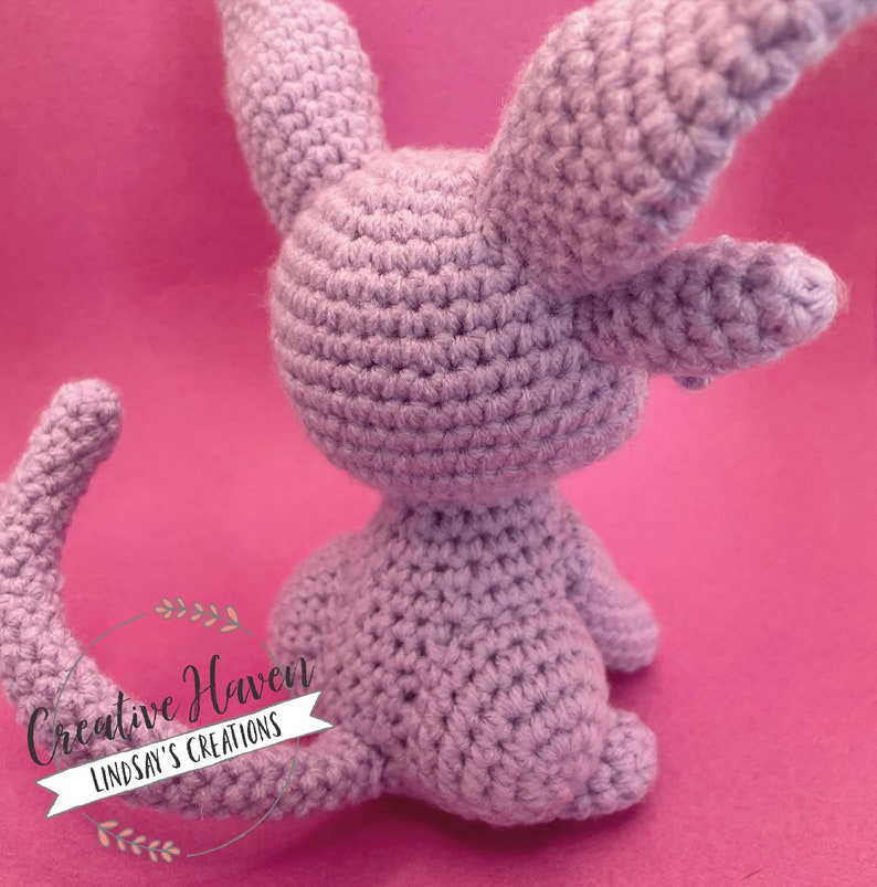 May include: A lavender-colored, crocheted bunny toy with long ears and a curved tail. The handmade plush animal is sitting on a pink background. The text "Creative Haven Lindsay's Creations" is in the lower left corner.