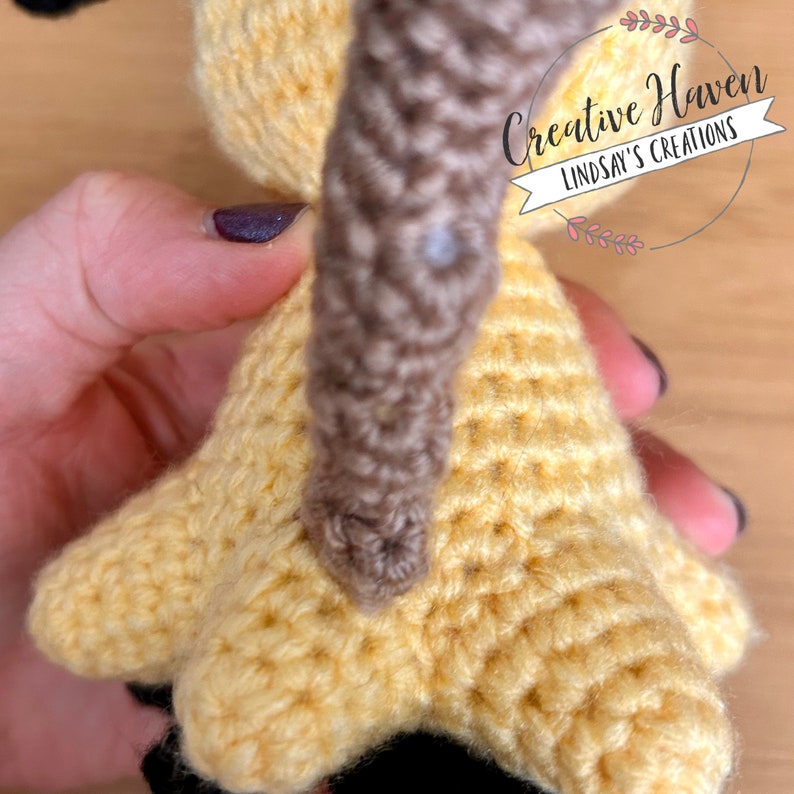 Puede incluir: Primer plano de un juguete amigurumi de crochet amarillo y marr&oacute;n. El juguete tiene una cola larga y marr&oacute;n y un cuerpo amarillo con una superficie texturizada. El juguete est&aacute; siendo sostenido por una mano.