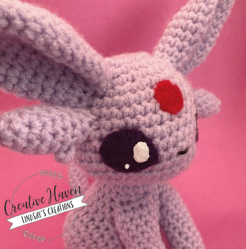 May include: A handmade crochet Espeon plushie in lavender yarn. The plushie features large ears, a red spot on its forehead, and dark purple eyes with white highlights. The text "Creative Haven Lindsay's Creations" is in the lower left corner.