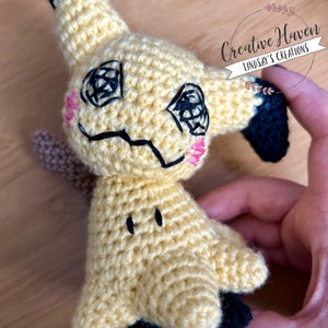 Puede incluir: Un juguete de peluche Pikachu amarillo tejido a crochet con orejas y cola negras. El juguete tiene ojos bordados, una boca negra y mejillas rosadas.
