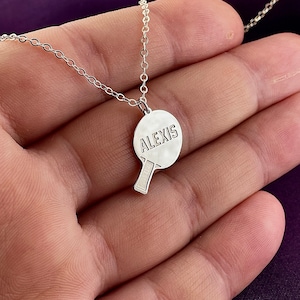 Puede incluir: Collar de plata con un colgante de raqueta de tenis de mesa. La raqueta está grabada con el nombre "Alexis".