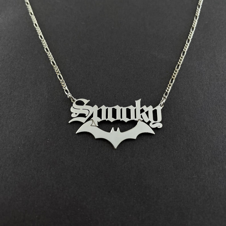 以下が含まれることがあります： ゴシック体で「Spooky」と書かれたバットペンダントが付いたシルバーネックレス