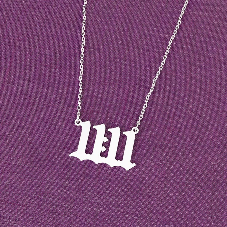 May include: A silver-colored necklace with the number 1111 in an old English font. The pendant hangs from a delicate chain against a purple background. The necklace is a piece of jewelry.