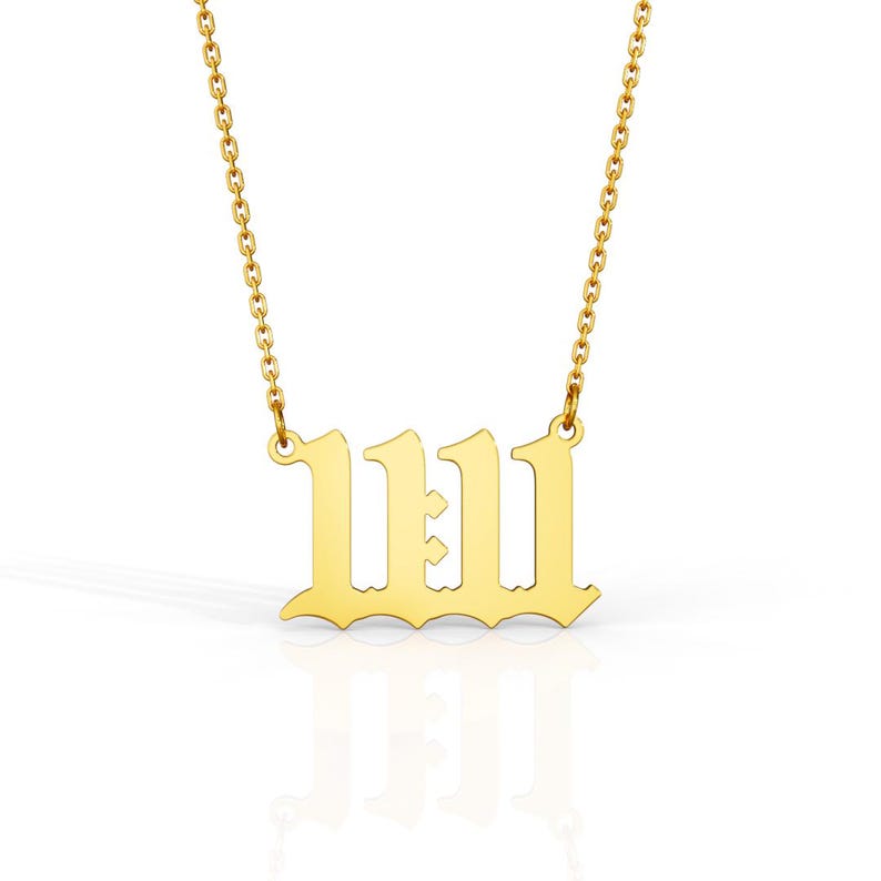May include: A gold-colored necklace with a chain and a pendant featuring the number 1111 in a gothic font. The pendant is centered and reflects light, set against a white background.