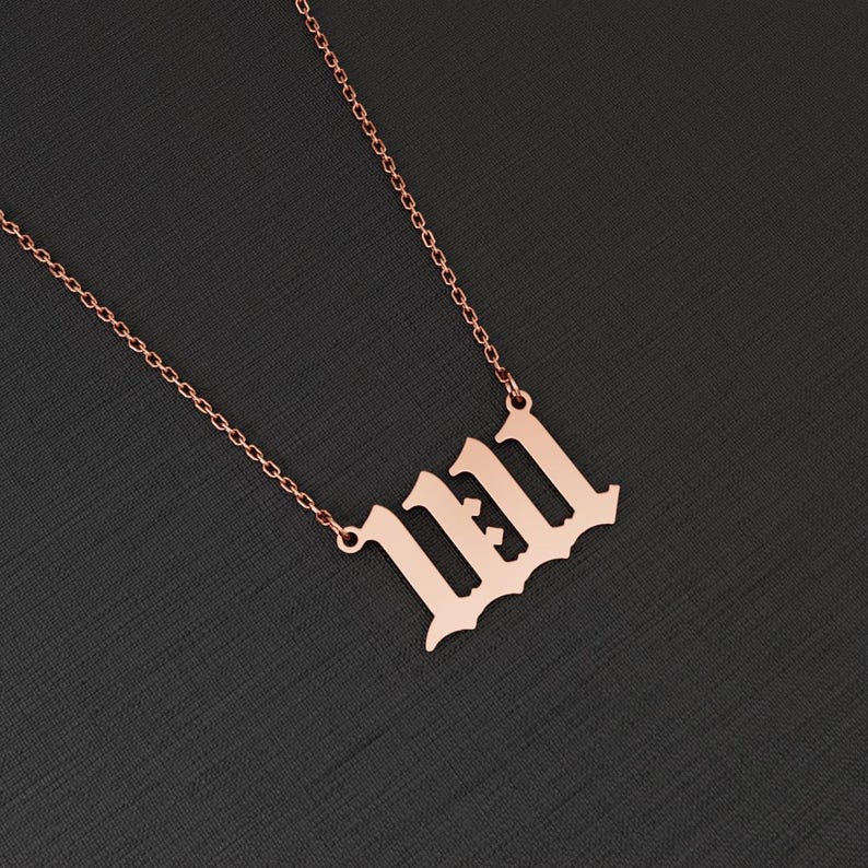 May include: Rose gold-colored necklace with the number 11:11 in an old English font. The pendant hangs from a delicate chain. The necklace is displayed against a dark gray background.