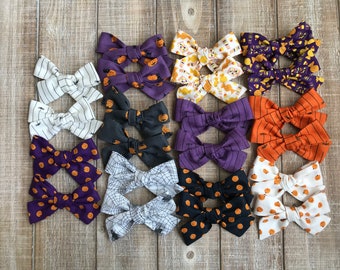 May include: A collection of Halloween-themed hair bows in various colors and patterns. Designs include pumpkins, stripes, spiderwebs, and candy corn. The bows are arranged on a wooden surface, showcasing a variety of festive styles.