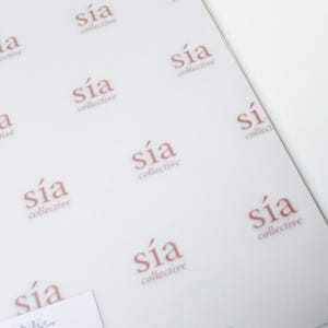 May include: White tissue paper featuring a repeating pattern of the words "sía collective" in a soft, reddish-brown hue. The paper is slightly translucent, and the design is evenly spaced. A small card with the words "THANK YOU!" is visible.