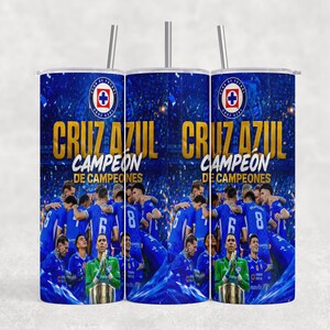 May include: A cylindrical tumbler with a metal straw featuring a blue graphic print of a soccer team celebrating on a field. The design includes the text Cruz Azul, Campeón de Campeones, and the team logo at the top against a stadium background.