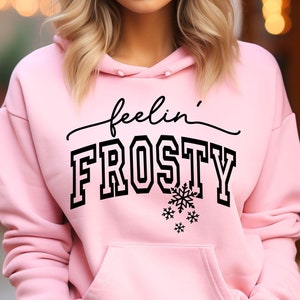 Puede incluir: Sudadera rosa con un gráfico negro que dice "feelin' FROSTY" con copos de nieve.