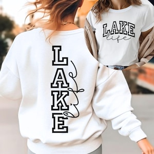 Può includere: Felpa bianca con la scritta "LAKE" stampata verticalmente in lettere nere sul retro. Un'immagine più piccola mostra una maglietta bianca con "LAKE life" in lettere nere.