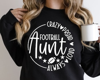May include: Black sweatshirt with the text "Crazy Proud Football Aunt Always" in a white circular design. The design includes a football, stars, and hearts. A white mug is held in the hand.