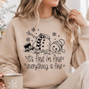 May include: A beige crewneck sweatshirt featuring a black graphic of a fallen snowman wearing a scarf and hat, surrounded by snowflakes. Below the illustration, the text reads: it's fine, i'm fine, everything is fine. The design is centered on the chest.