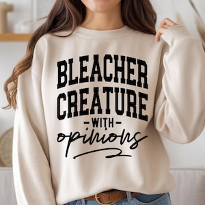 May include: A beige sweatshirt with the text "BLEACHER CREATURE - WITH - opinions" in black. The sweatshirt has a crew neck and long sleeves. The text is in a bold, blocky font with the word "opinions" in a cursive font.