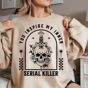 May include: A beige sweatshirt with a black graphic design. The design features a skull with a dagger, roses, and the text "YOU INSPIRE MY INNER SERIAL KILLER". The sweatshirt has a crew neck and long sleeves.
