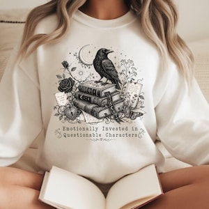 May include: A cream-colored crewneck sweatshirt featuring a black ink-style graphic of a raven perched on a stack of three books. The design includes roses, a crescent moon, a heart-shaped lock, and the text "Emotionally Invested in Questionable Characters."