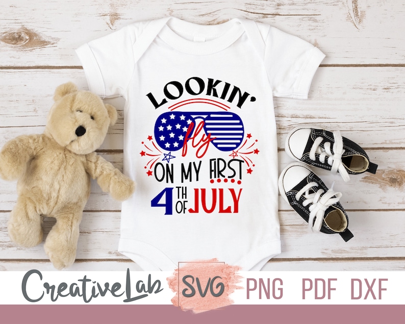 May include: White baby bodysuit with the text "Lookin' Fly on my First 4th of July" in red, white, and blue. The design includes sunglasses with a star-filled lens. A teddy bear and black and white baby shoes are next to the bodysuit.