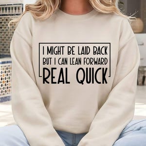 以下が含まれることがあります： クリーム色のスウェットシャツ。黒い長方形の中に「I MIGHT BE LAID BACK BUT I CAN LEAN FORWARD REAL QUICK」という黒い文字がプリントされています。カジュアルなスタイルで、普段使いに適しています。