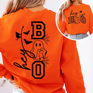 Puede incluir: Sudadera naranja con un dise&ntilde;o negro con tem&aacute;tica de Halloween. El dise&ntilde;o incluye las palabras "hey BOO" con un fantasma, murci&eacute;lagos y telara&ntilde;as. La sudadera es de cuello redondo y manga larga.