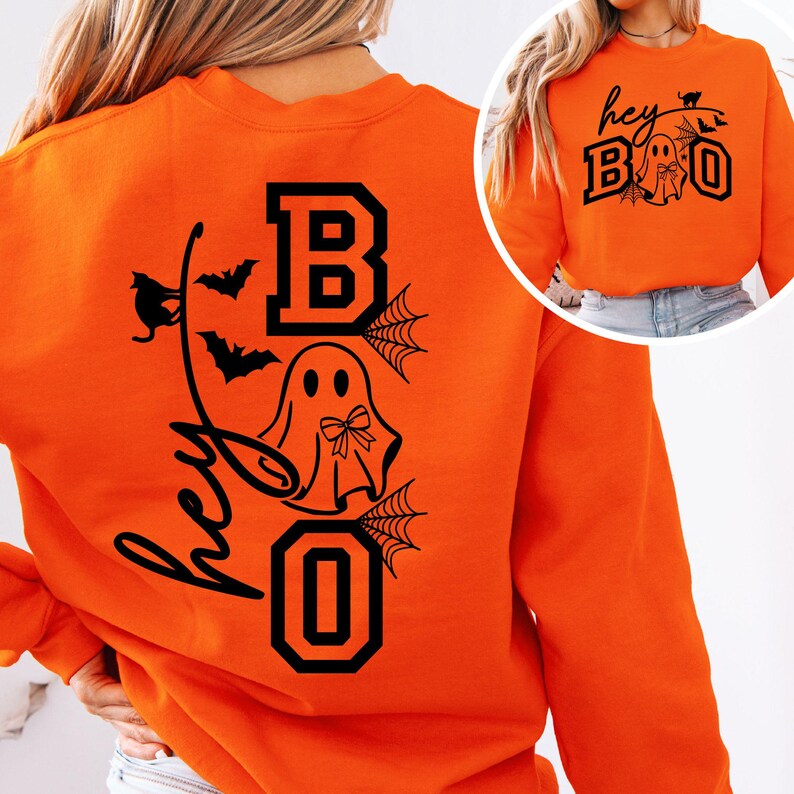 Puede incluir: Sudadera naranja con un dise&ntilde;o negro con tem&aacute;tica de Halloween. El dise&ntilde;o incluye las palabras "hey BOO" con un fantasma, murci&eacute;lagos y telara&ntilde;as. La sudadera es de cuello redondo y manga larga.