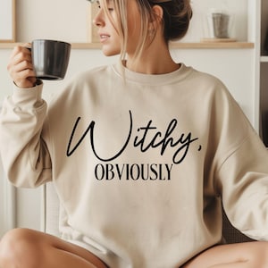 May include: A beige crewneck sweatshirt featuring the black text Witchy, OBVIOUSLY printed on the front. The garment has long sleeves and a ribbed neckline, providing a simple and clean look for casual wear in a neutral tone.