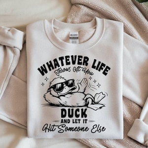 May include: Cream-colored sweatshirt with a black graphic of a duck wearing sunglasses, with the text "WHATEVER LIFE Throws at You DUCK AND LET IT Hit Someone Else."
