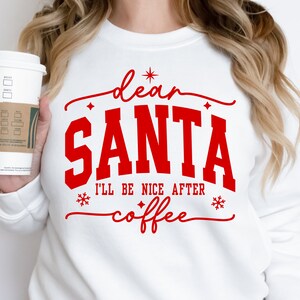 May include: White sweatshirt with the text "dear SANTA I'LL BE NICE AFTER coffee" in red. The design includes snowflakes and star accents. A person is holding a white coffee cup with a brown sleeve.