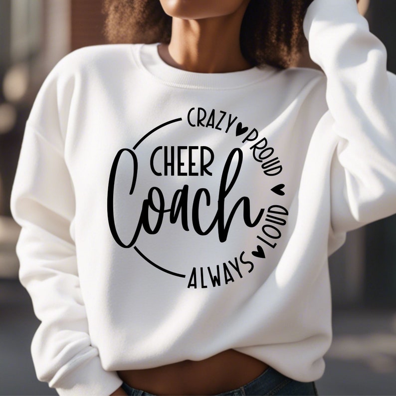 May include: White sweatshirt featuring the text "CHEER Coach" in a black circular design. The words "CRAZY", "PROUD", "LOUD", and "ALWAYS" are also printed in black around the circle, with heart symbols. A casual, comfortable top.