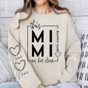 Puede incluir: Una sudadera beige con el texto "this MIMI wears her on her sleeve" en un cuadro negro con un corazón en el lado derecho. El texto "Name" está escrito dos veces en la manga izquierda en forma de corazón.