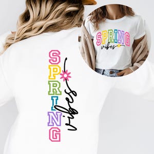 May include: White t-shirt with the word "SPRING" in rainbow colours and the words "vibes" in black script. The back of the shirt has the word "SPRING" vertically in rainbow colours with a flower and the words "vibes" in black script.